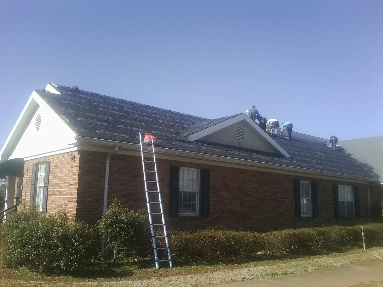 Crew tearing shingles off a brick ranch roof with a ladder set — Emergency Roof Repair work by our crew in Beecher