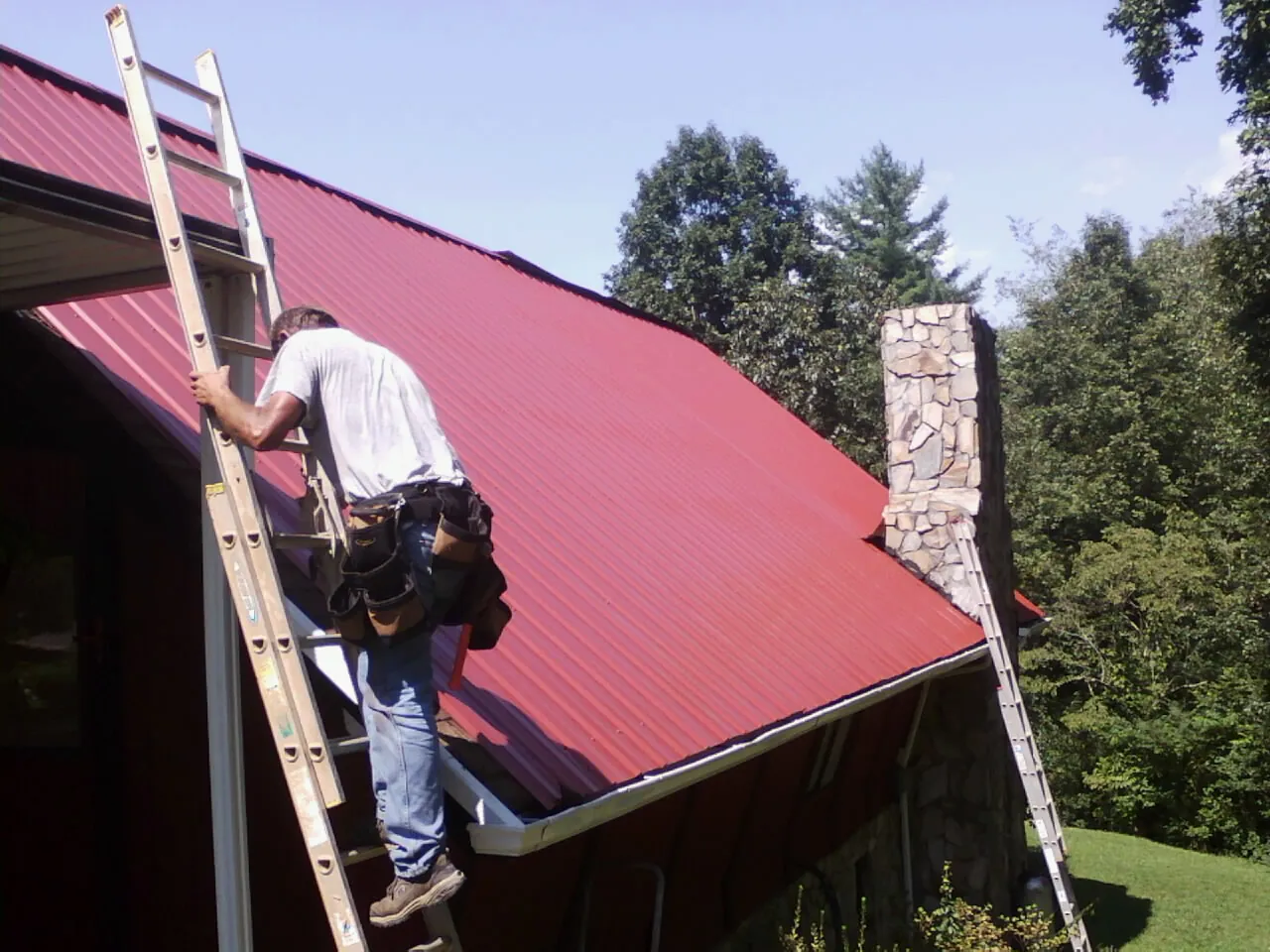 Roofer on a ladder beside a red metal roof and stone chimney — Soffit & Fascia Repair by Roofing Brothers in Beecher